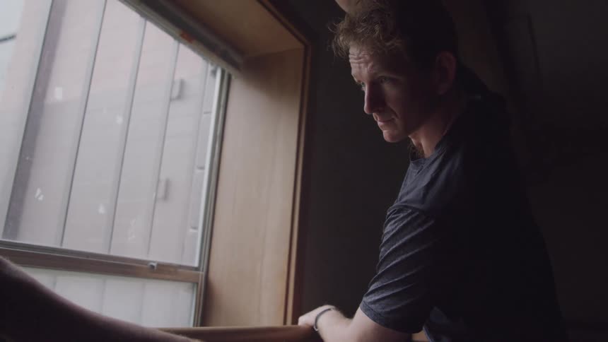 A man is performing a ballet stretch with one leg extended on a barre in front of a window illuminated by light streaming through the window