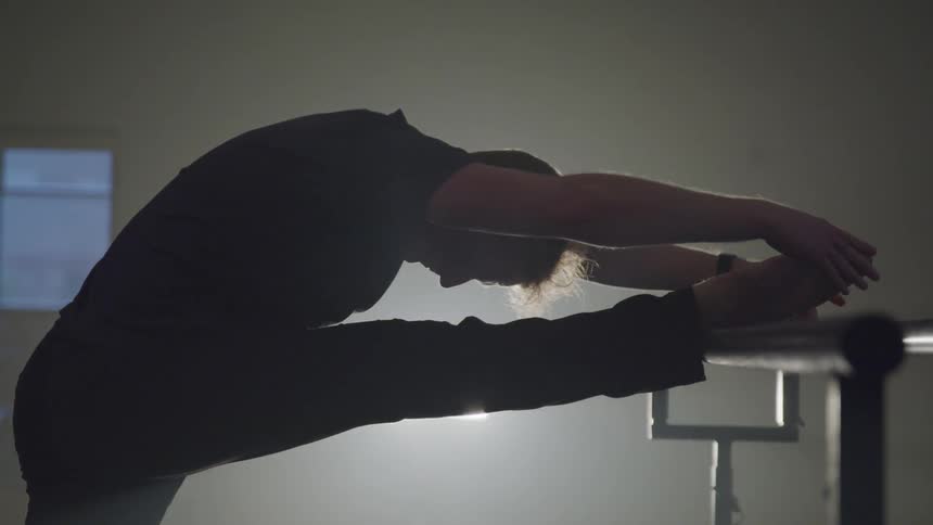 A person is performing a stretching exercise with one leg extended on a ballet barre in a dimly lit dance studio