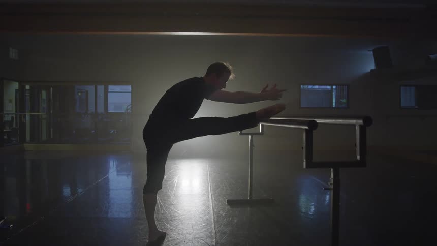 A person is performing a stretching exercise with one leg extended on a ballet barre in a dimly lit dance studio