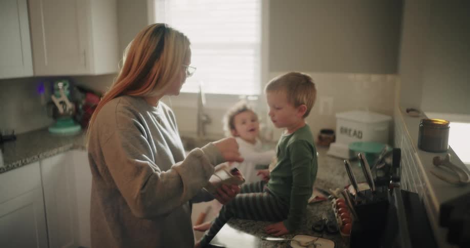A woman in a gray sweater stands in a kitchen preparing food with two young children nearby