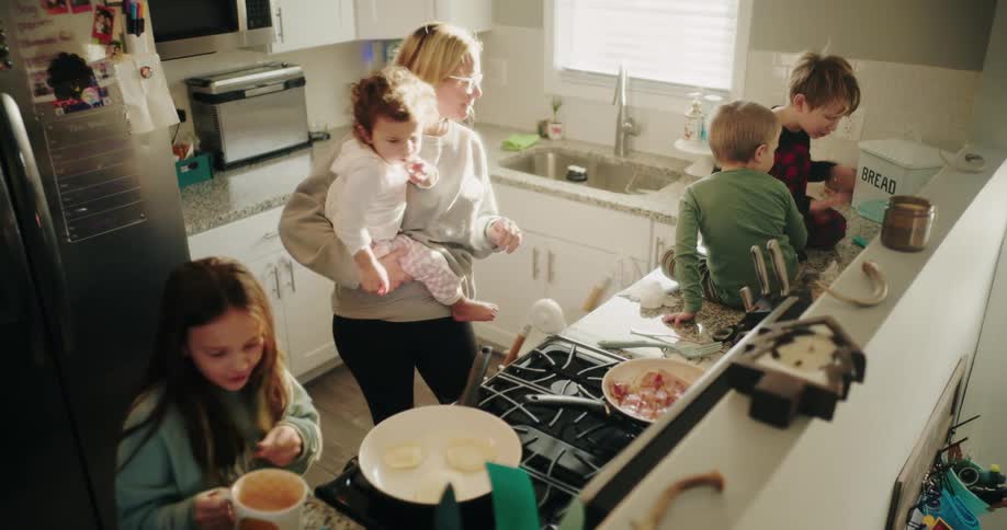 A mother holds a baby and cooks in a busy kitchen with three other children