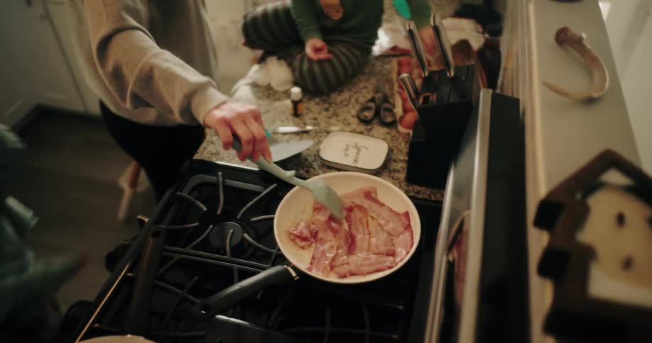 Adult hand with spatula frying bacon in frying pan and accompanied by children
