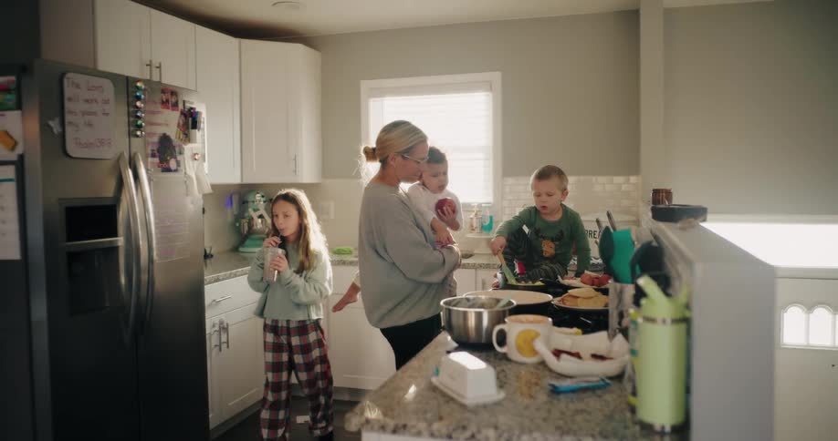 A mother holds a baby while overseeing her children in a busy kitchen at mealtime