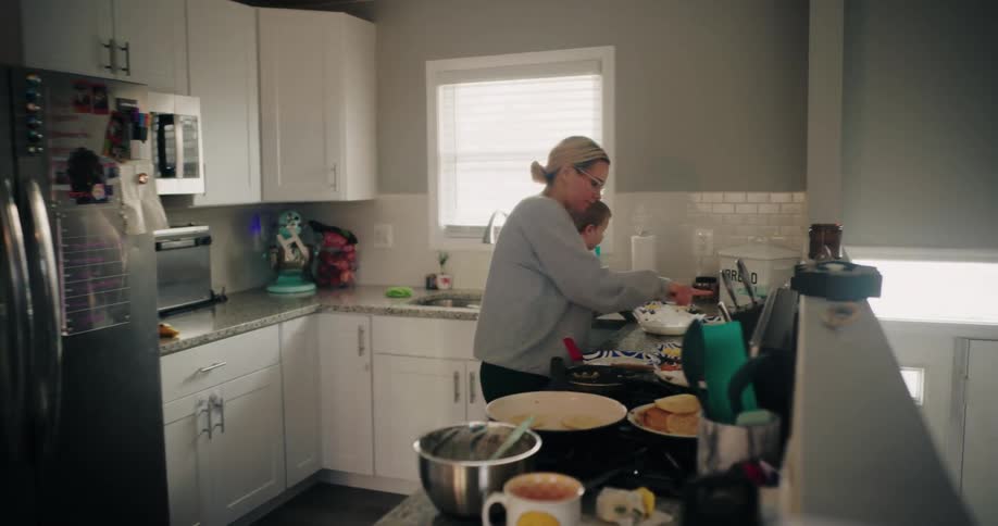 In a brightly lit slightly messy kitchen a woman cooks while a young child watches from the counter