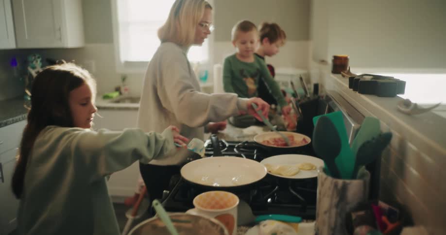 Mother in the kitchen frying bacon next to her young children