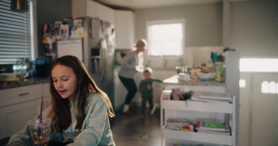 A kitchen scene with a child sitting at a table with a drink and a mother playing with another child while preparing food in the background