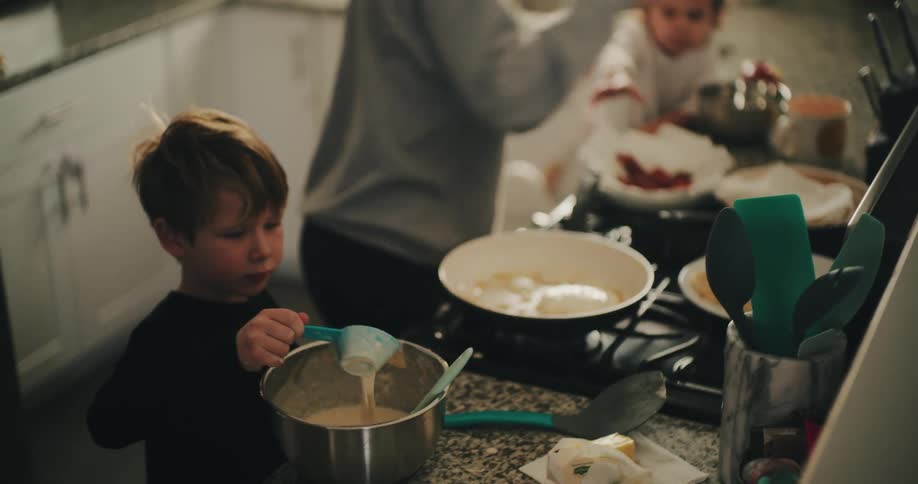 A young boy stirs batter in a bowl as a woman cooks pancakes and a baby plays in the background