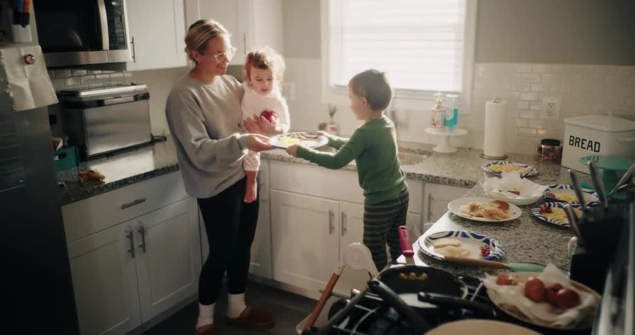 Mother with a child on her lap handing a plate to her other small child in the kitchen