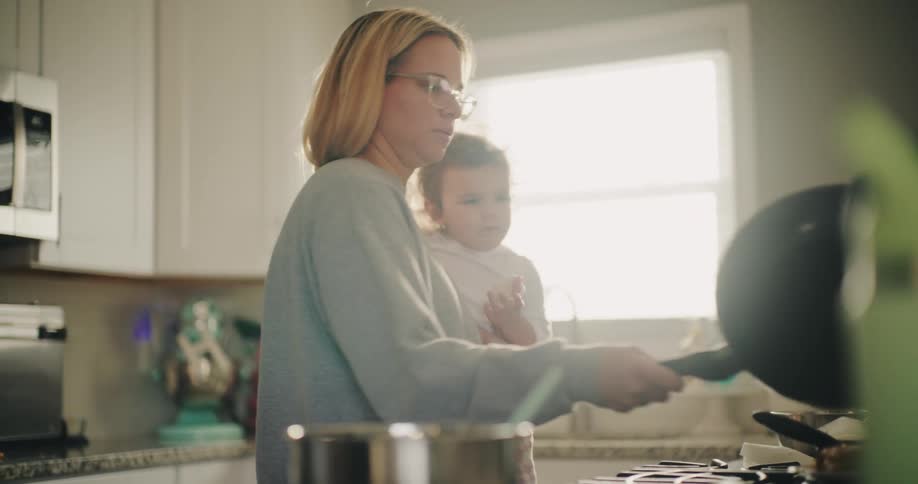 A woman with glasses holds a baby while cooking in a kitchen