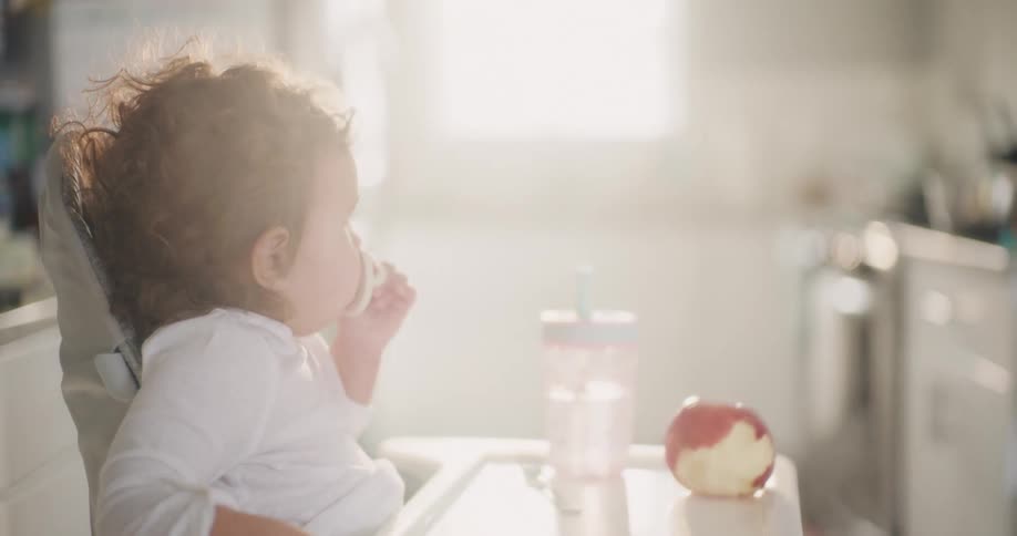 Toddler sucking on a pacifier sits in a highchair in a brightly lit kitchen