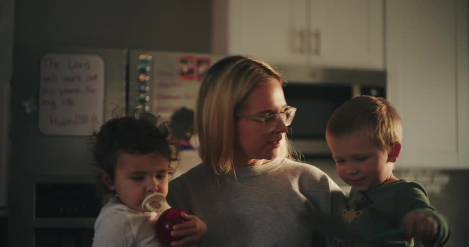 Mother holds two young children in the kitchen while her son holds a spatula