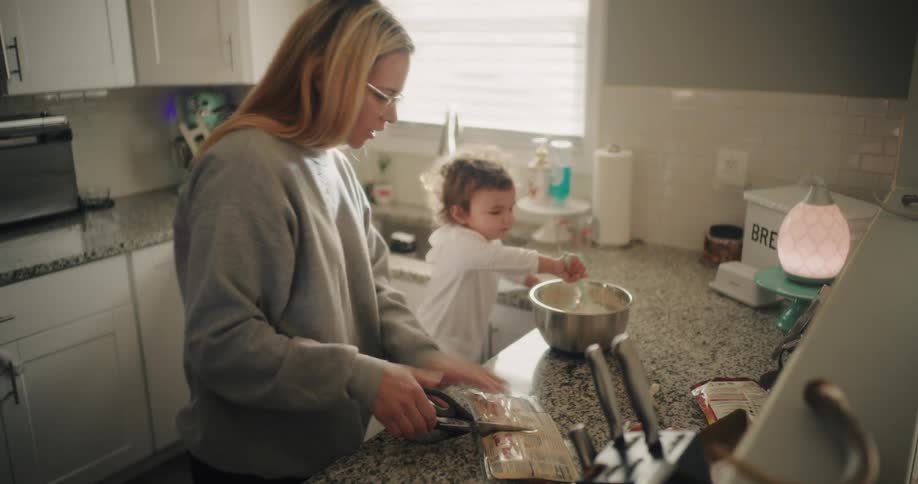 A mother is cutting open a package while a child mixes ingredients in a large bowl on the kitchen countertop