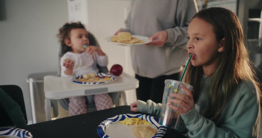 A little girl is eating something from a cup with a straw and there is a little baby girl who has a spoon in her mouth looking