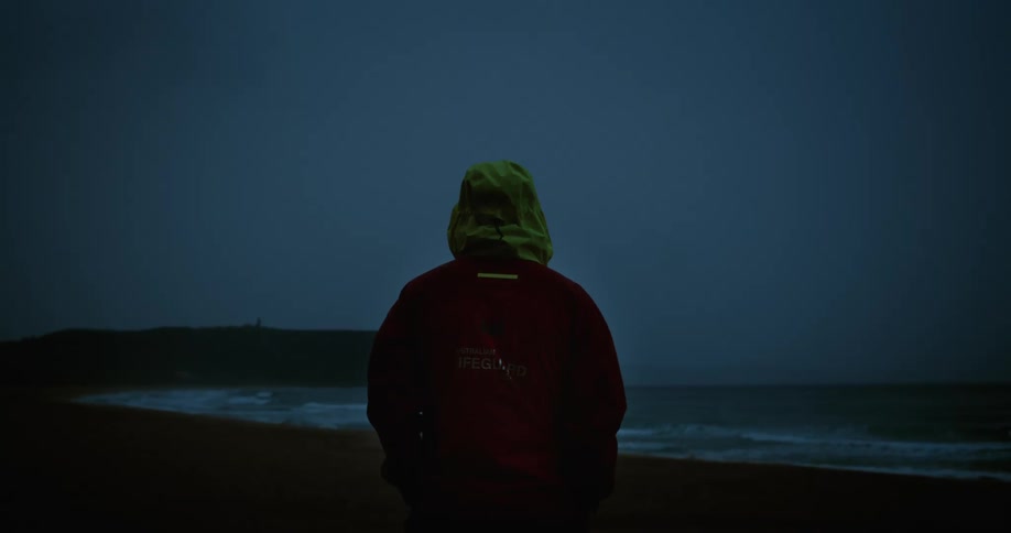 A hooded person in a jacket stands on a beach as waves break on the shore under a dark sky