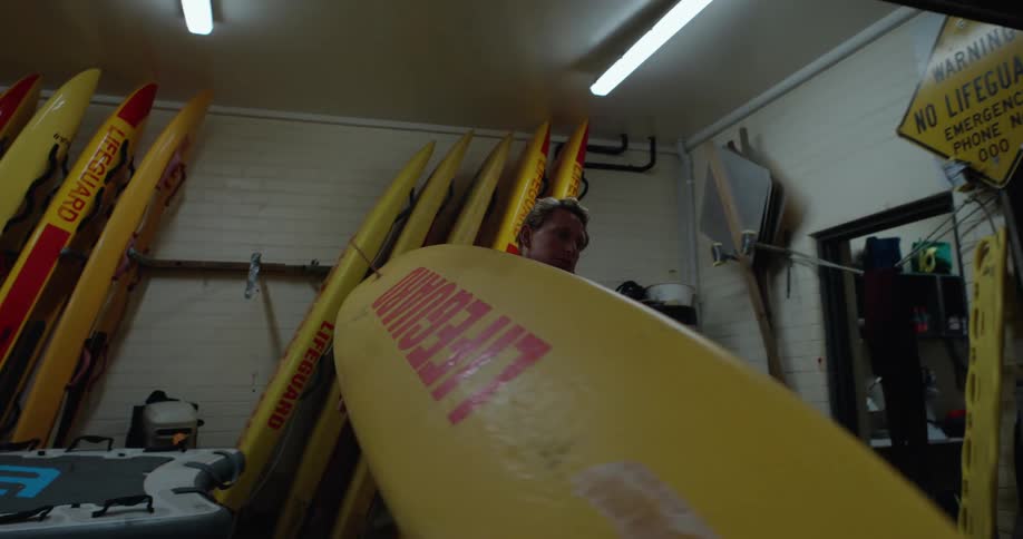 A lifeguard in a red outfit is carrying a yellow surfboard in a storage room filled with multiple yellow surfboards