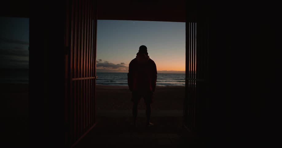 A person is standing in the doorway of a dark structure silhouetted against a sunset over the ocean
