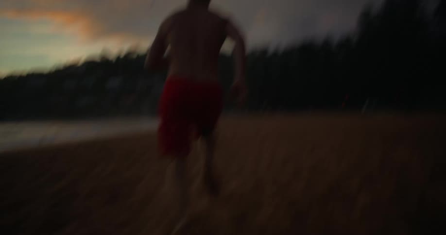 Shirtless man in red shorts running on sand at dawn