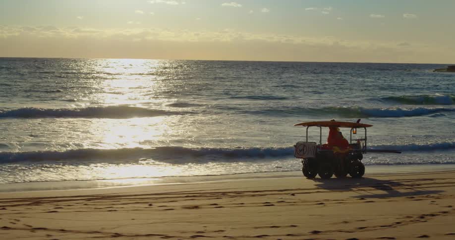 A lifeguard vehicle is driving along a sandy beach at sunset with the sun reflecting off the ocean waves in the background