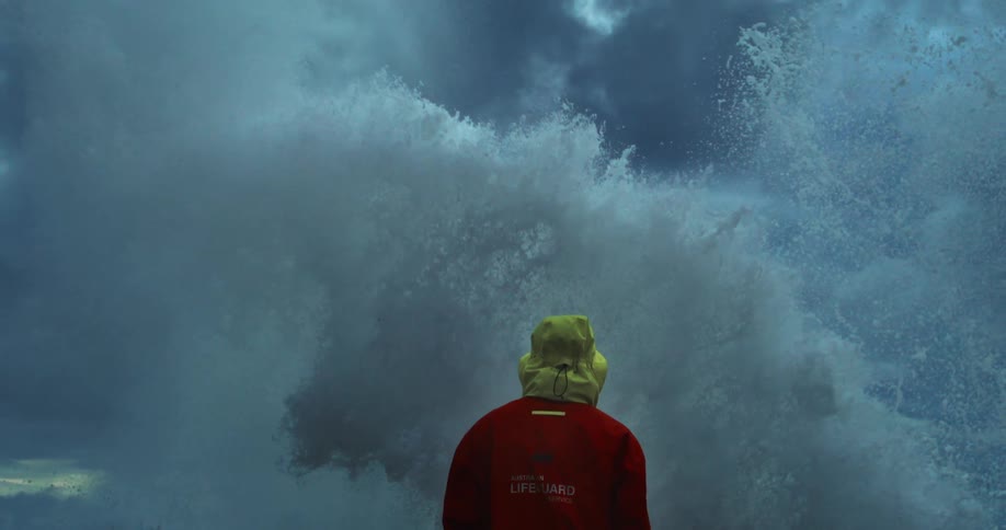 A lifeguard in a red jacket and yellow hood observes a massive wave crashing against the coast