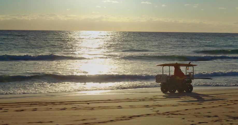 A cart with two people inside driving along the edge of a beach with some waves breaking on the shore