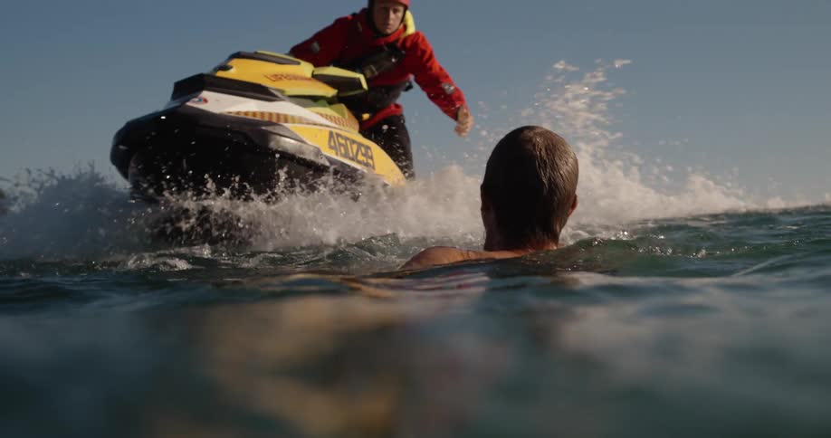 A person in a red jacket is helping a shirtless person out of the water onto a green and white jet ski with a distant shoreline visible in the background