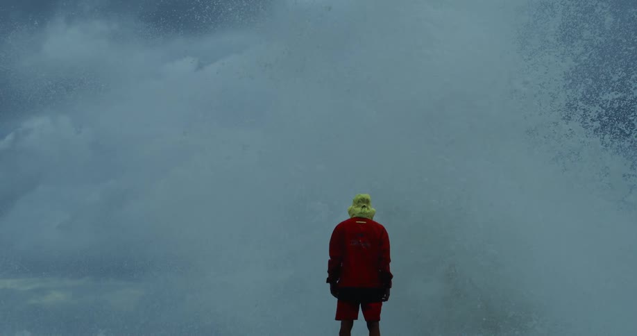 A man wearing a red jacket and yellow headgear stands facing a massive ocean wave crashing against the shore under a dark stormy sky