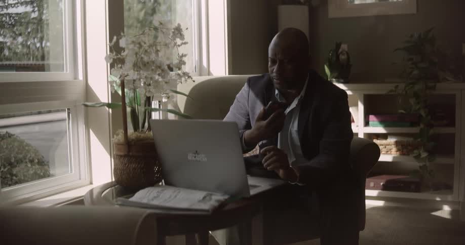 A person is sitting in a chair by a window working on a laptop with a potted plant and some documents on the table