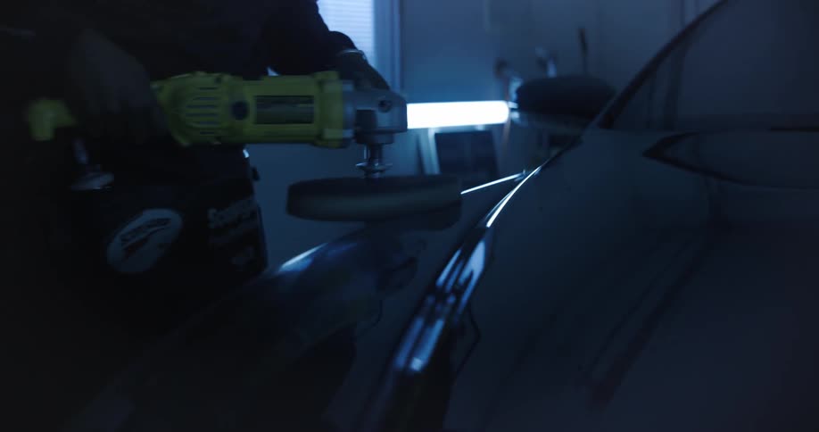 A man is using a power tool to polish the surface of a car in a garage or workshop setting