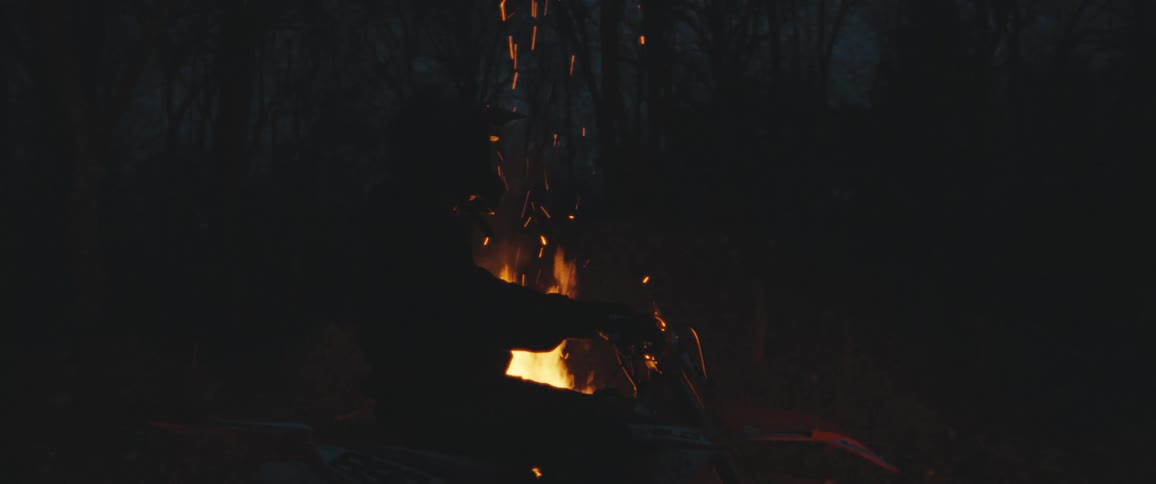 A person is sitting on a motorcycle in front of a campfire at night with sparks flying upwards and trees silhouetted in the background