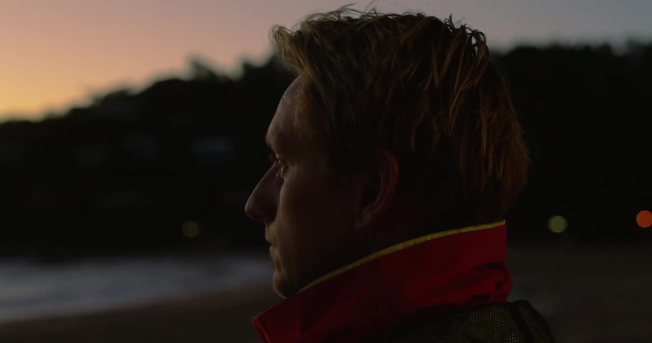 The face of a man wearing a red jacket looking at the ocean at sunset