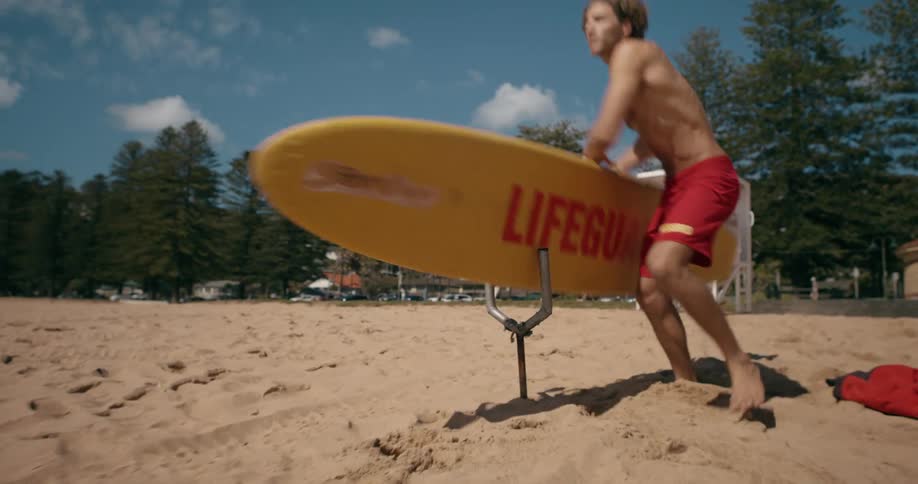 A lifeguard is running to get a yellow surfboard that is on the sand and throws his clothes on the ground