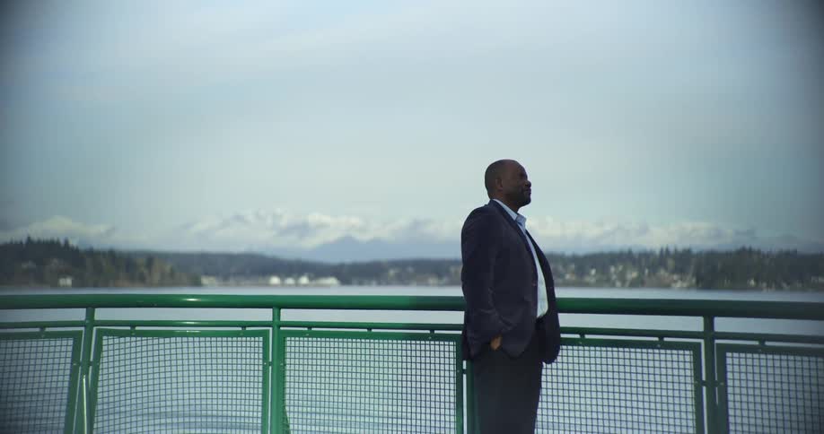 Man in gray suit outdoors by a lake with a city in the background