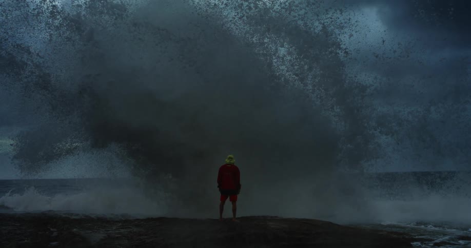 A lifeguard in a red jacket and yellow hat is momentarily engulfed by a giant wave crashing ashore on a rocky coast