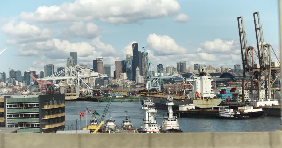 A bustling port with several cargo ships cranes and tugboats set against a city skyline with highrise buildings and a partly cloudy sky