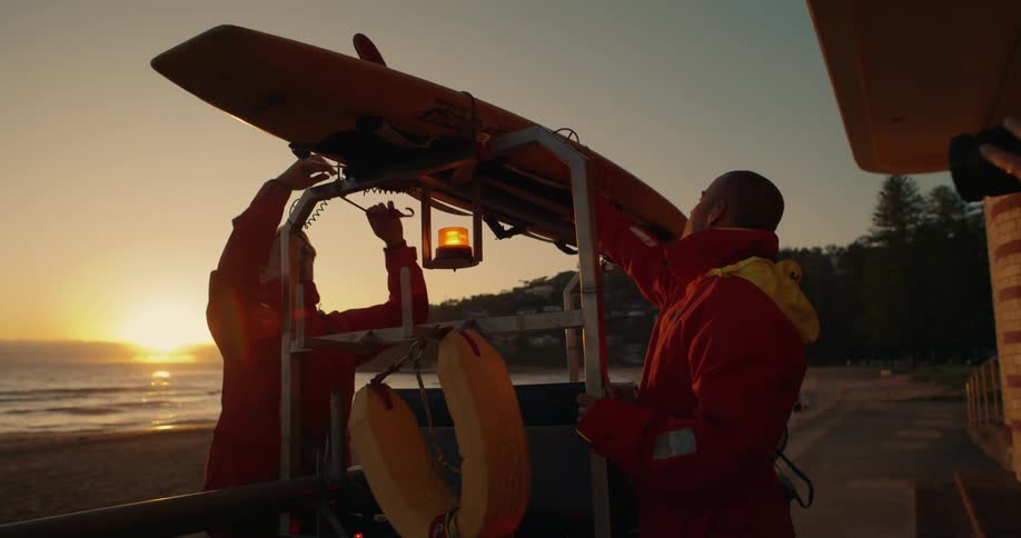 A couple of lifeguards are tying a surfboard to the roof of a small vehicle on the coast at sunset