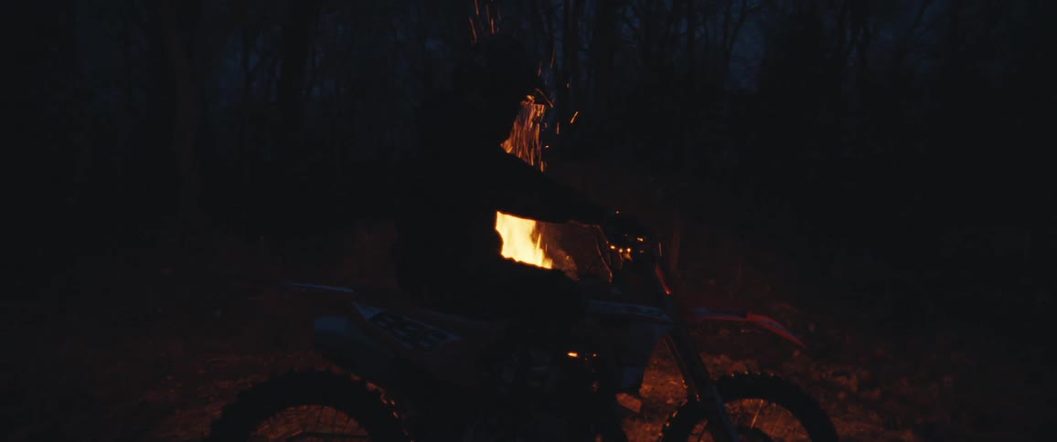 Silhouette of a motorcyclist standing at night in front of a campfire in the middle of the forest