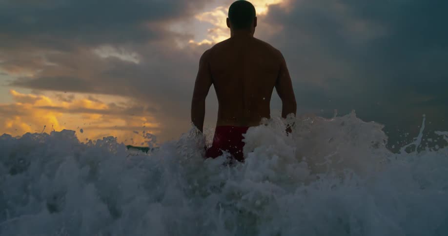 A person is standing waistdeep in the ocean facing away from the camera with waves crashing around and a dramatic sunset sky in the background