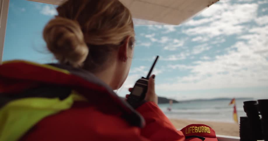 A lifeguard in a red jacket is using a walkietalkie while looking out at a beach