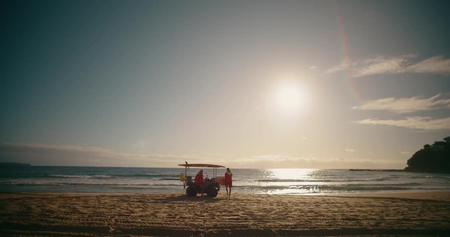 A small car is parked on a sandy beach near the shoreline with two lifeguards walking out of it the sun shining brightly over the ocean and a clear sky above
