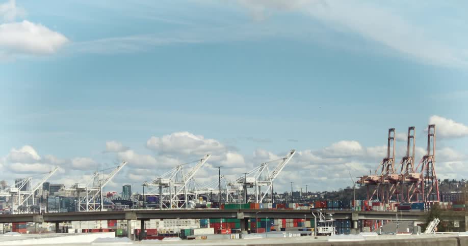 A wide shot captures numerous cranes and shipping containers in a port setting under a blue sky dotted with white clouds with a cityscape visible in the distance