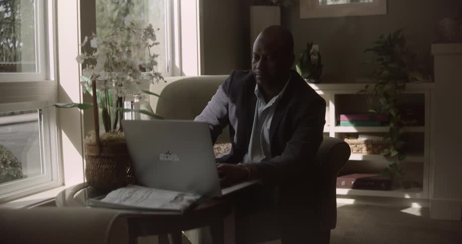 An adult man wearing formal wear sitting in a living room using a laptop near a window