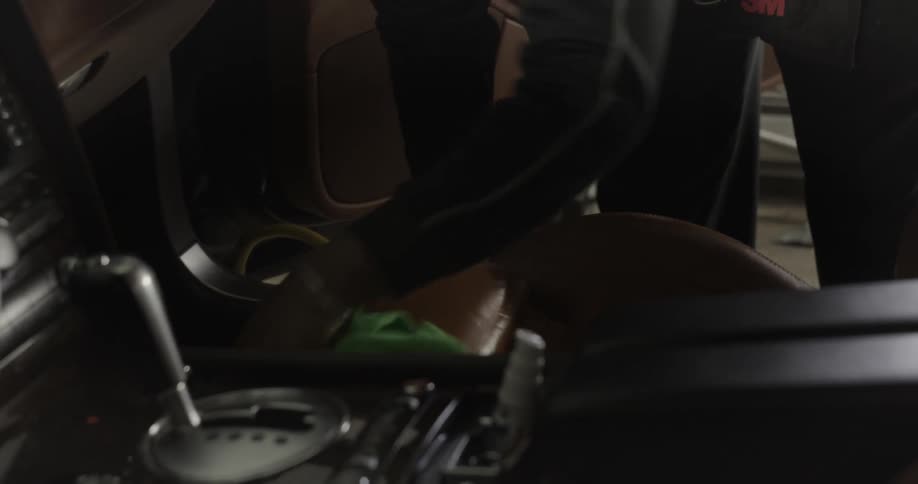 A person is cleaning the interior of a car near the drivers seat with a green cloth and various cleaning supplies are visible