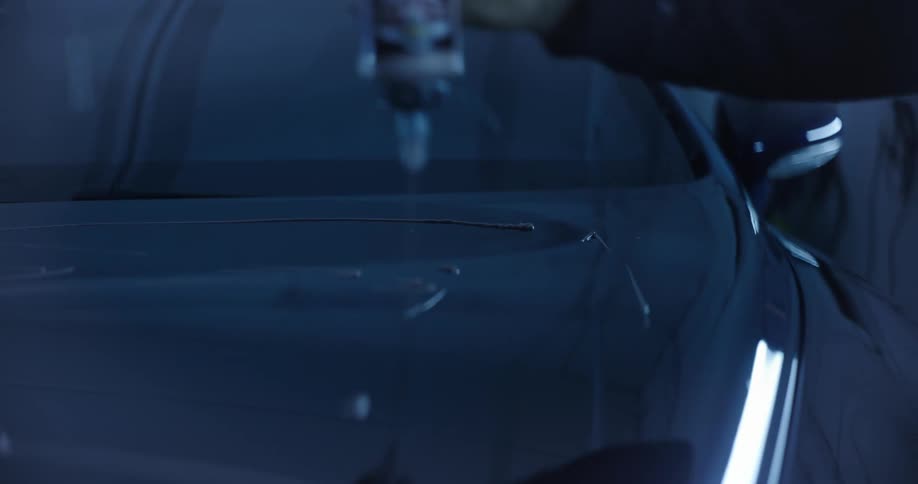 A person applies a polishing liquid from a bottle onto the hood of a car in a dimly lit garage