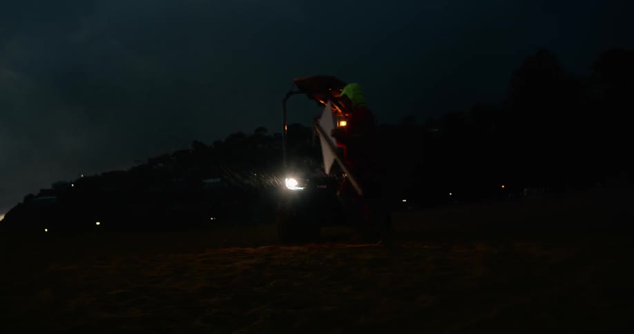 A car parked on a beach with headlights on at night and a person is placing a flag in the sand while it is raining