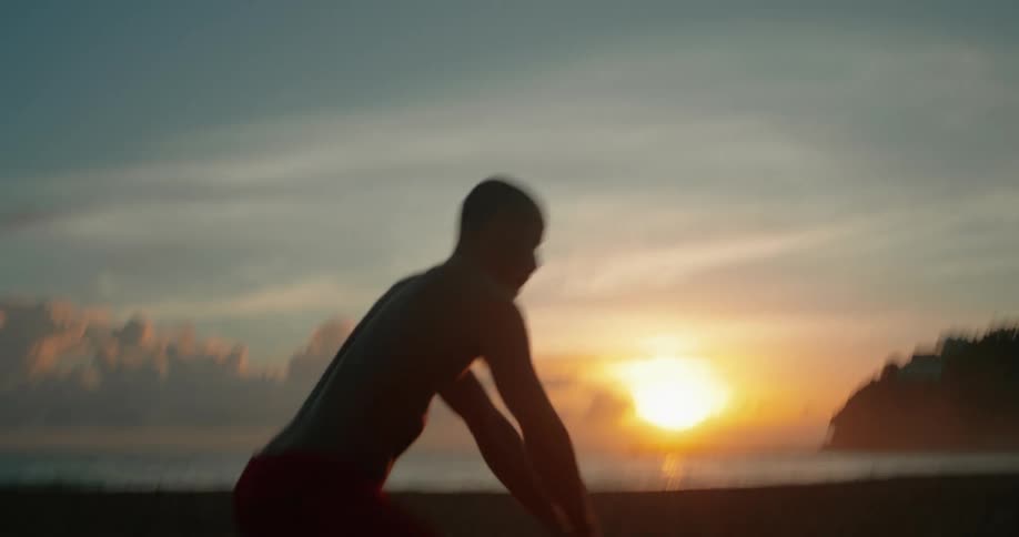 A man on a beach is lifting a kettlebell swinging it from between his legs to over his head the sun is rising behind him