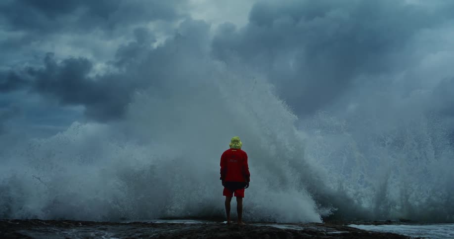 A person in a red outfit and yellow hat stands on a rocky shore facing a massive wave crashing against the rocks under a stormy sky
