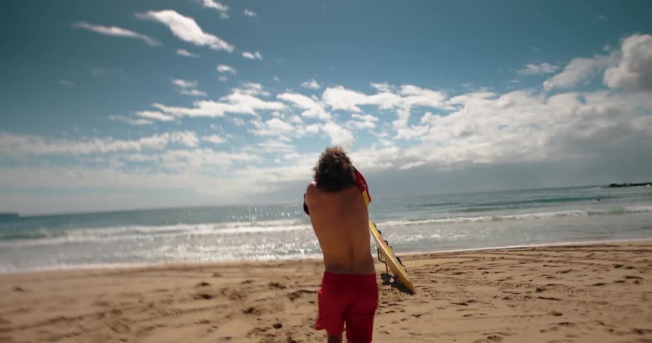 A lifeguard runs on the beach taking off his jacket and picking up a surfboard