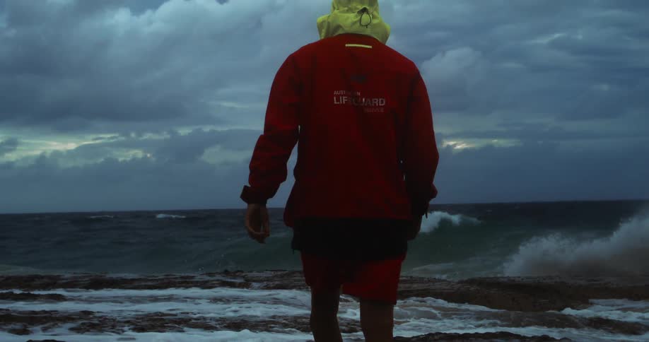 Lifeguard in a red jacket walks toward the rough sea under a cloudy sky