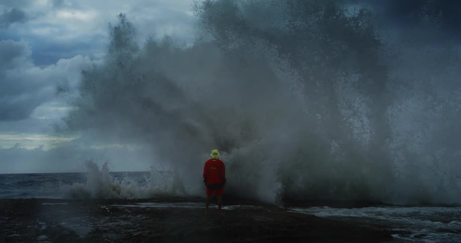A person in red jacket and yellow hat stands on the shore as a massive wave crashes engulfing them in water