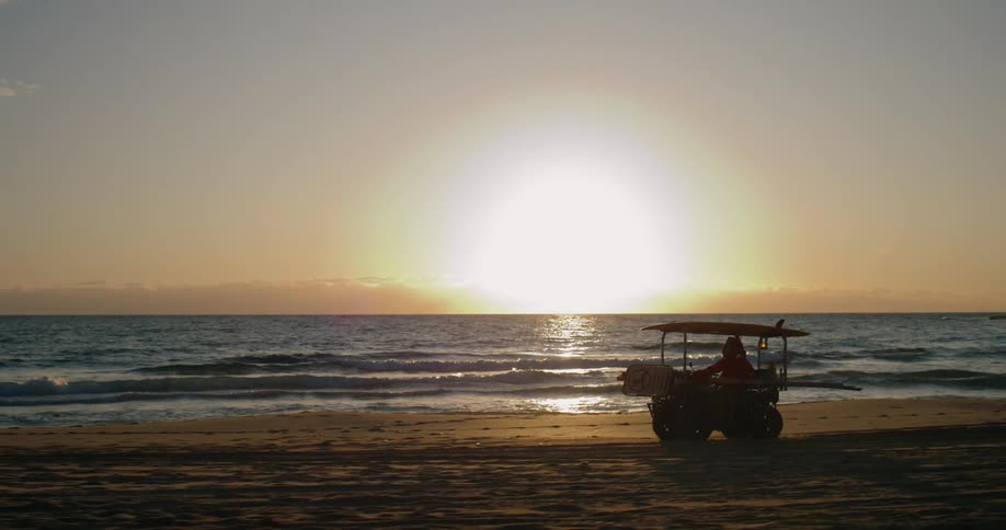 A lifeguard vehicle is driving along a sandy beach at sunset with the sun reflecting off the ocean waves in the background